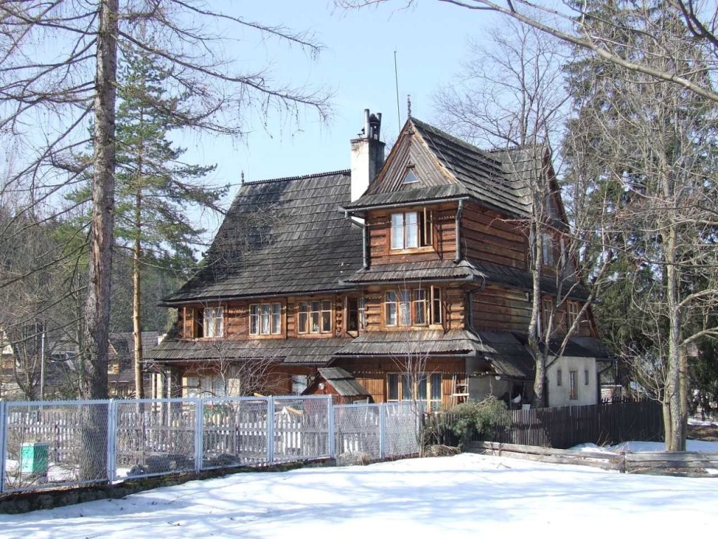 Na zdjęciu duży, drewniany dom otoczony śniegiem i drzewami. Dach jest dwuspadowy, o zmiennym nachyleniu, z dużym kominem po środku. Wygląd domu nawiązuje do tradycyjnego budownictwa góralskiego, tzw. stylu zakopiańskiego. 