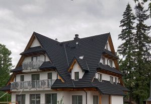 Zdjęcie przedstawia duży, biały dom z czarnym dachem. Dom jest dwupiętrowy, z wysokim spadzistym dachem sięgającym aż do parteru. Na każdym piętrze umieszczone są balkony z żelaznymi balustradami. Fasada domu jest pomalowana na biało. 