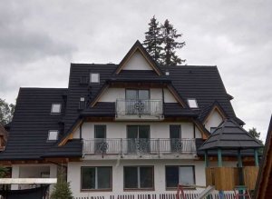 Zdjęcie ukazuje duży, biały dom z czarną dachówką. Dom jest dwupiętrowy, z wysokim spadzistym dachem sięgającym aż do parteru. Na każdym piętrze umieszczone są balkony z żelaznymi balustradami. Fasada domu jest pomalowana na biało. 