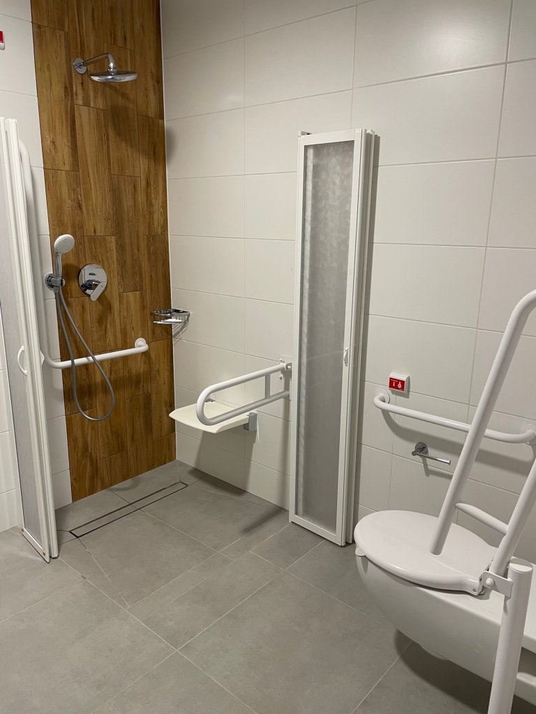 Wnętrze łazienki z dostosowaniami dla osób z niepełnosprawnościami. Na wprost w kącie prostopadłych ścian umieszczony jest prysznic. Pionowy pas, na którym zawieszona jest wylewka prysznicowa oraz bateria, wyłożony jest kafelkami w kolorze ciemnego drewna. Obok, na prostopadłej ścianie powieszone jest białe krzesełko prysznicowe. Obszar prysznica wydzielają białe drzwiczki ze szklanymi wypełnieniami, rozkładane w harmonijkę. Część po prawej stronie jeszcze nie została powieszona – drzwiczki opierają się o ścianę. Po lewej stronie kadru częściowo widoczny jest sedes z podniesionym uchwytem obok. Ściany łazienki wyłożone są białymi kafelkami, podłogę wykładają kafle w kolorze szarym.