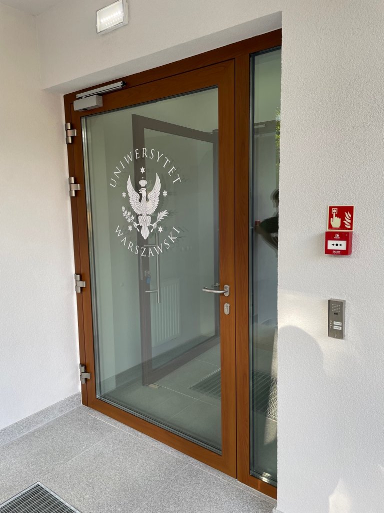 Drzwi wejściowe do budynku. Drzwi są szerokie, w drewnianej stolarce, z dużym przeszkleniem na całej wysokości. Na przeszkleniu umieszczone jest godło uczelni otoczone jego nazwą „Uniwersytet Warszawski”. Drzwi otwierane są na zewnątrz, w lewo.