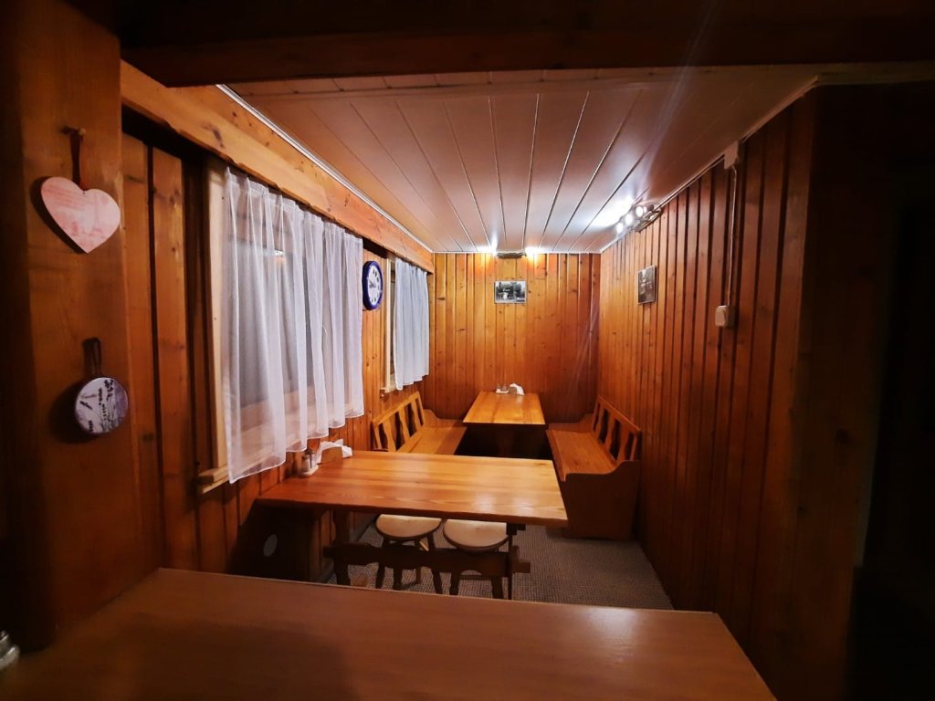 Na zdjęciu część jadalniana w której znajdują się dwa stoły drewniane z dwoma ławkami oraz dwa taborety drewniane. Całość wykończona boazerią drewnianą, po lewej stronie dwa okna z widokiem na ogród.