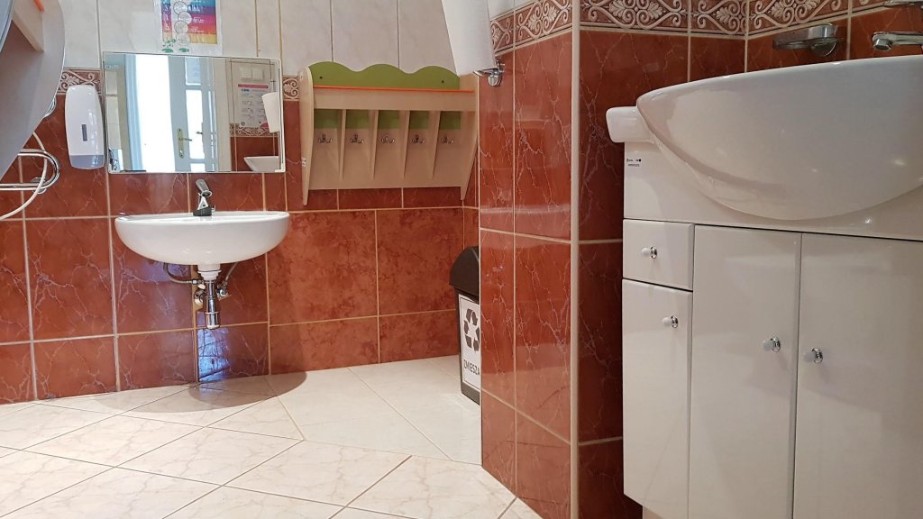 Zdjęcie przedstawia wnętrze łazienki. Po lewej stronie znajduje się umywalka dla dzieci z wiszącym lustrem. Na ścianie wyłożonej kafelkami wiszą półki z wieszaczkami na ręczniki . Po prawej stronie umywalka wraz z szafką.