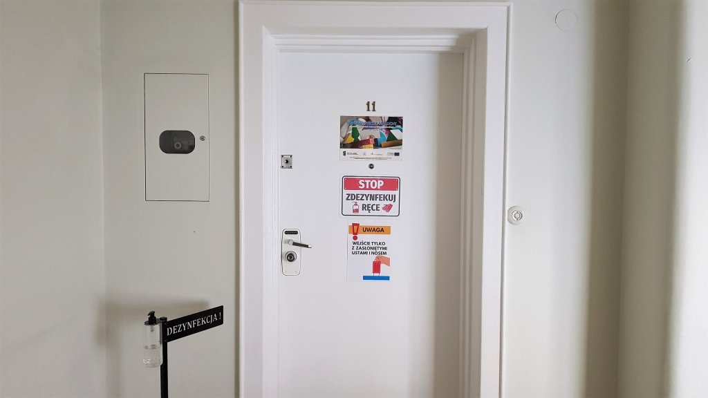 Zdjęcie drzwi wejściowych do lokalu żłobka. Na białych drewnianych drzwiach wiszą tabliczki informacyjne. Po lewej stronie stojak z płynem do dezynfekcji rąk.