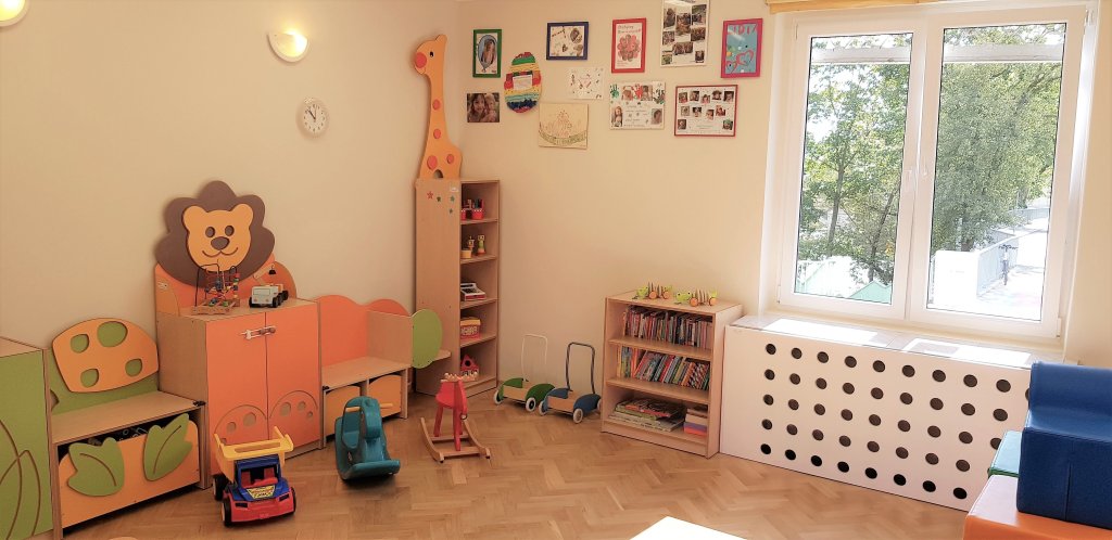 Zdjęcie przedstawia wnętrze sali zabaw. Dziecięce meble mają kształt lwa i żyrafy. Na półkach i drewnianej podłodze stoją zabawki. Ściany ozdobione są zdjęciami i laurkami. Przez okno widać drzewo z zielonymi liśćmi.