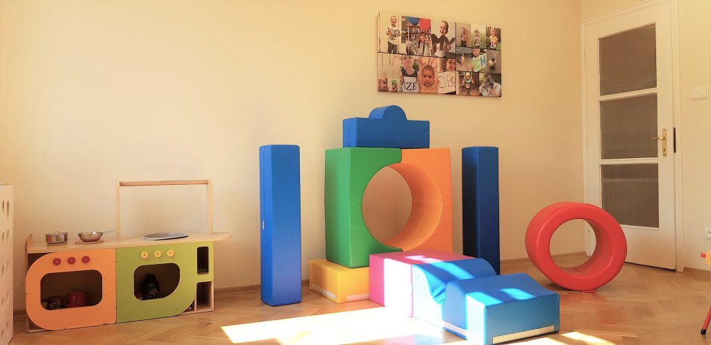 Zdjęcie przedstawia wnętrze sali zabaw z innej perspektywy. Na drewnianej podłodze poukładane są kolorowe pianki rehabilitacyjne, w rogu po lewej stronie dziecięca kuchenka . Na ścianie wisi obraz ze zdjęć dzieci. 
