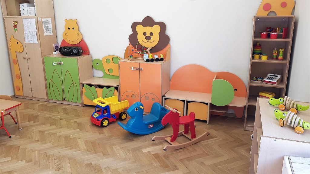 Zdjęcie przedstawia wnętrze sali zabaw z innej perspektywy. Dziecięce meble mają kształt żyrafy, hipopotama, lwa, żółwia i lwa. Na półkach poukładane są zabawki. Na drewnianej podłodze stoją koniki na biegunach oraz samochód ciężarowy. 