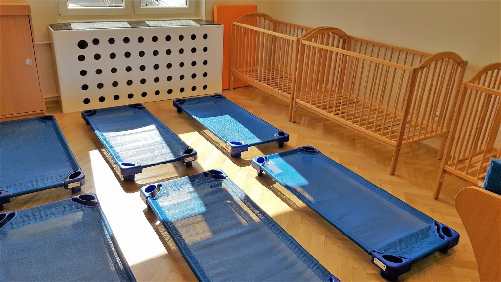 Zdjęcie Przedstawia wnętrze sypialni. Na drewnianej podłodze stoją leżaczki dla dzieci, przy ścianie widać drewniane łóżeczka szczebelkowe.