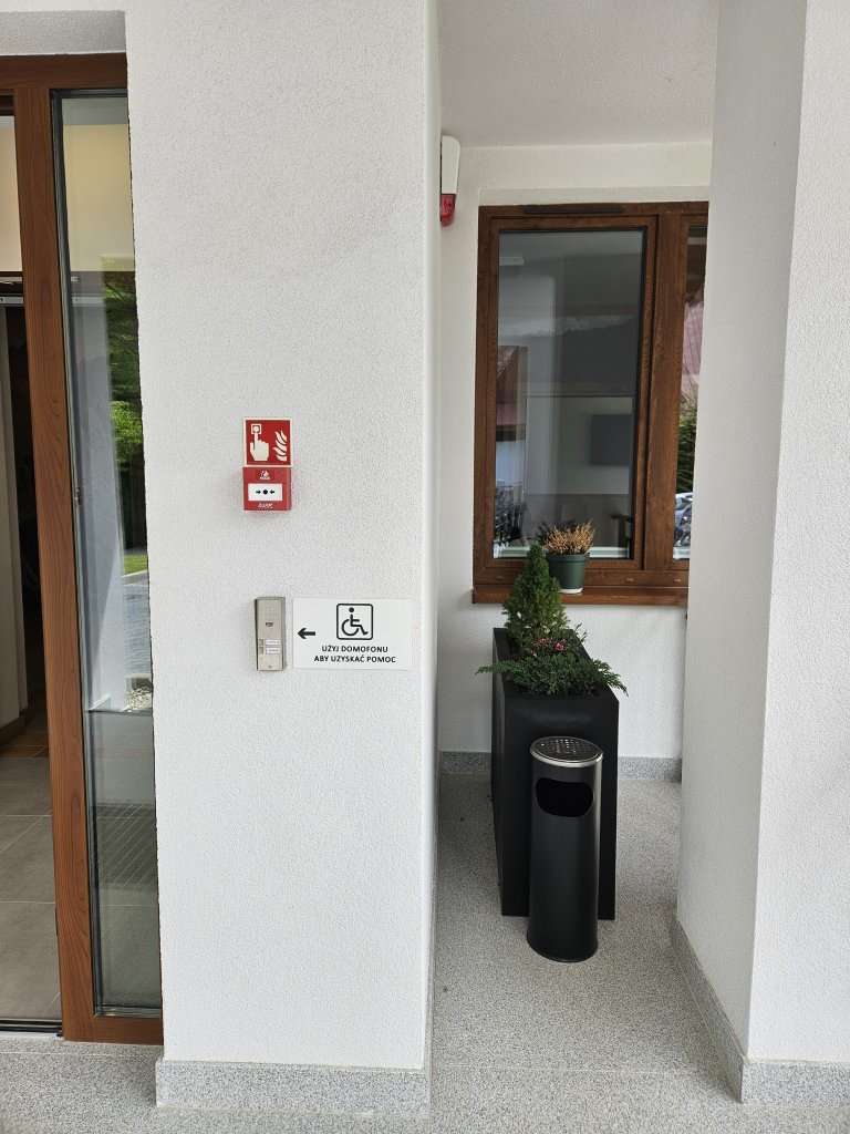 Zdjęcie przedstawia wejście do budynku, na wprost domofon, nad nim przycisk alarmu pożarowego II stopnia ROP, po prawej stronie donica z kwiatami i okno