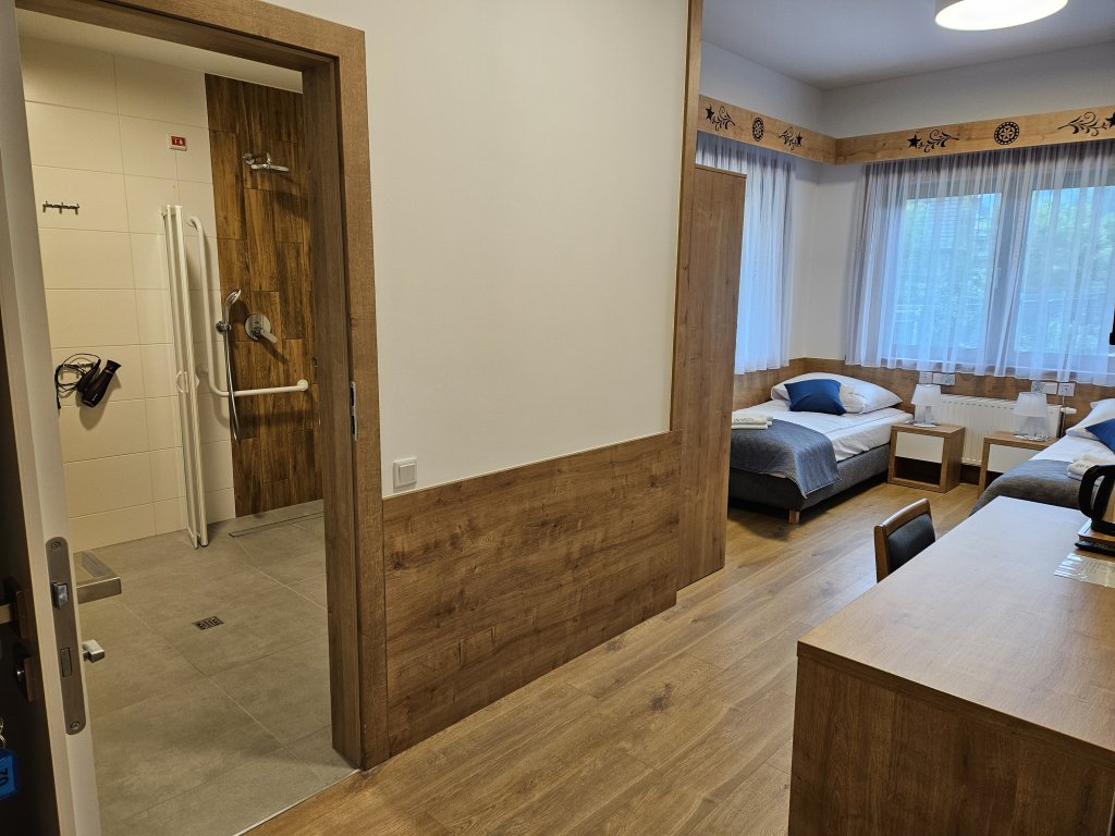 Pokój nr 2 dla osób niepełnosprawnych: po lewej widać łazienkę z prysznicem i suszarką, w centrum - ściana , w głębi - okna i dwa osobne łóżka, z prawej z przodu - biurko.